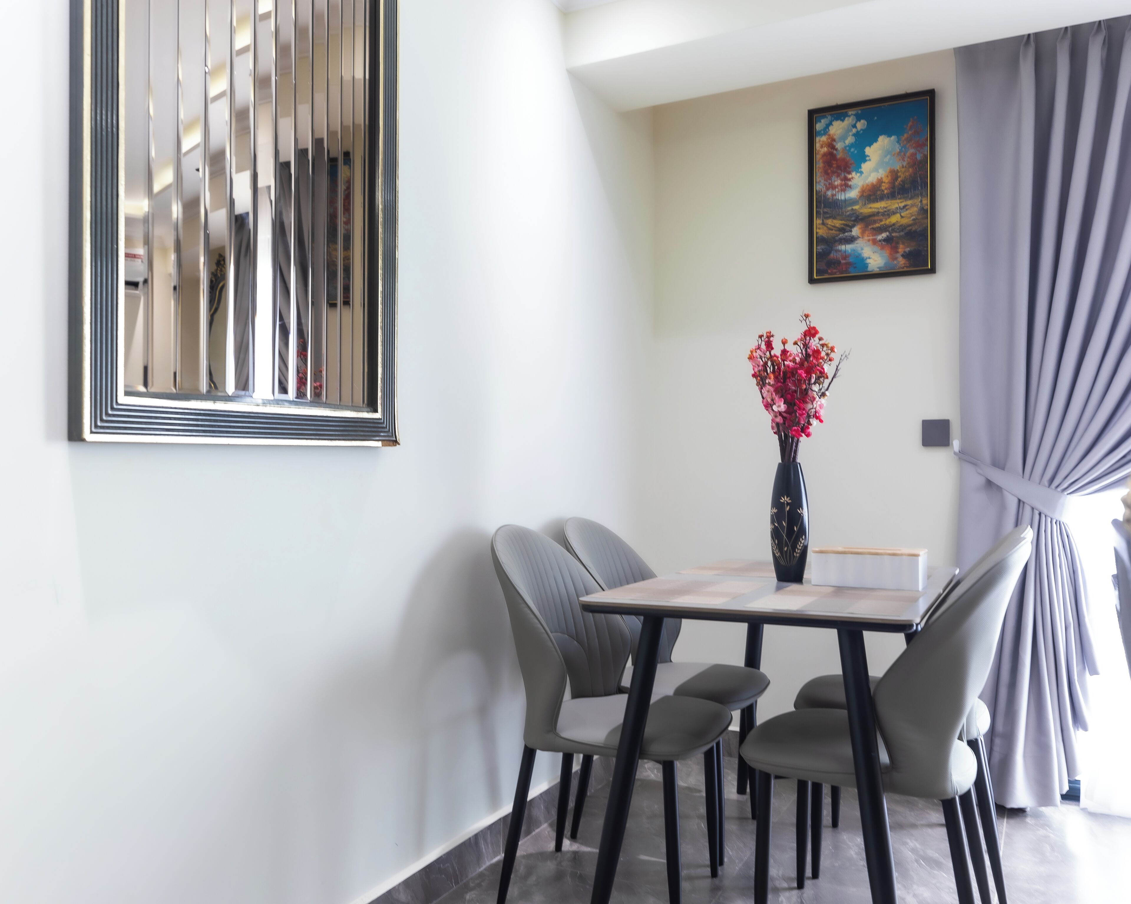 Un comedor con una mesa, sillas, un florero con flores, un cuadro en la pared y una ventana con cortinas.