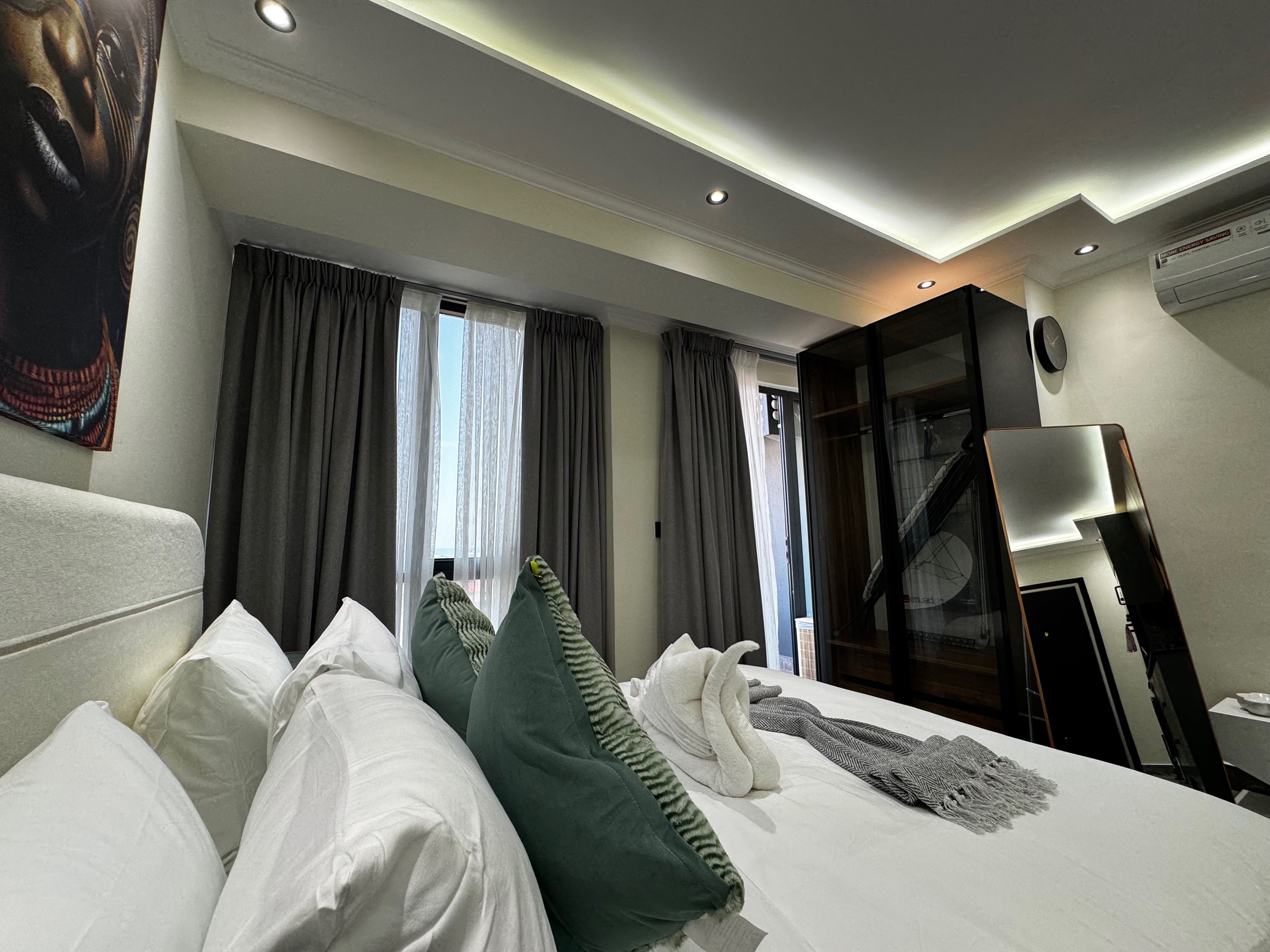 A hotel room with a bed, pillows, a large window with curtains, a wardrobe, and a mirror.