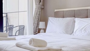 Una cama bien hecha con ropa blanca, un cabecero con paneles grises, una lámpara blanca sobre una mesita de noche y una silla junto a la ventana.
