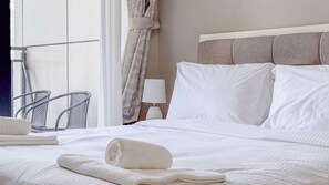 Ein ordentlich bezogenes Bett mit weißen Leinen, ein grauer Kopfteil, eine weiße Lampe auf einem Nachttisch und ein Stuhl am Fenster.