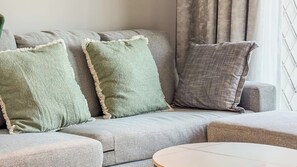 Un sofá gris con tres almohadones, una mesa redonda y una ventana con cortinas.