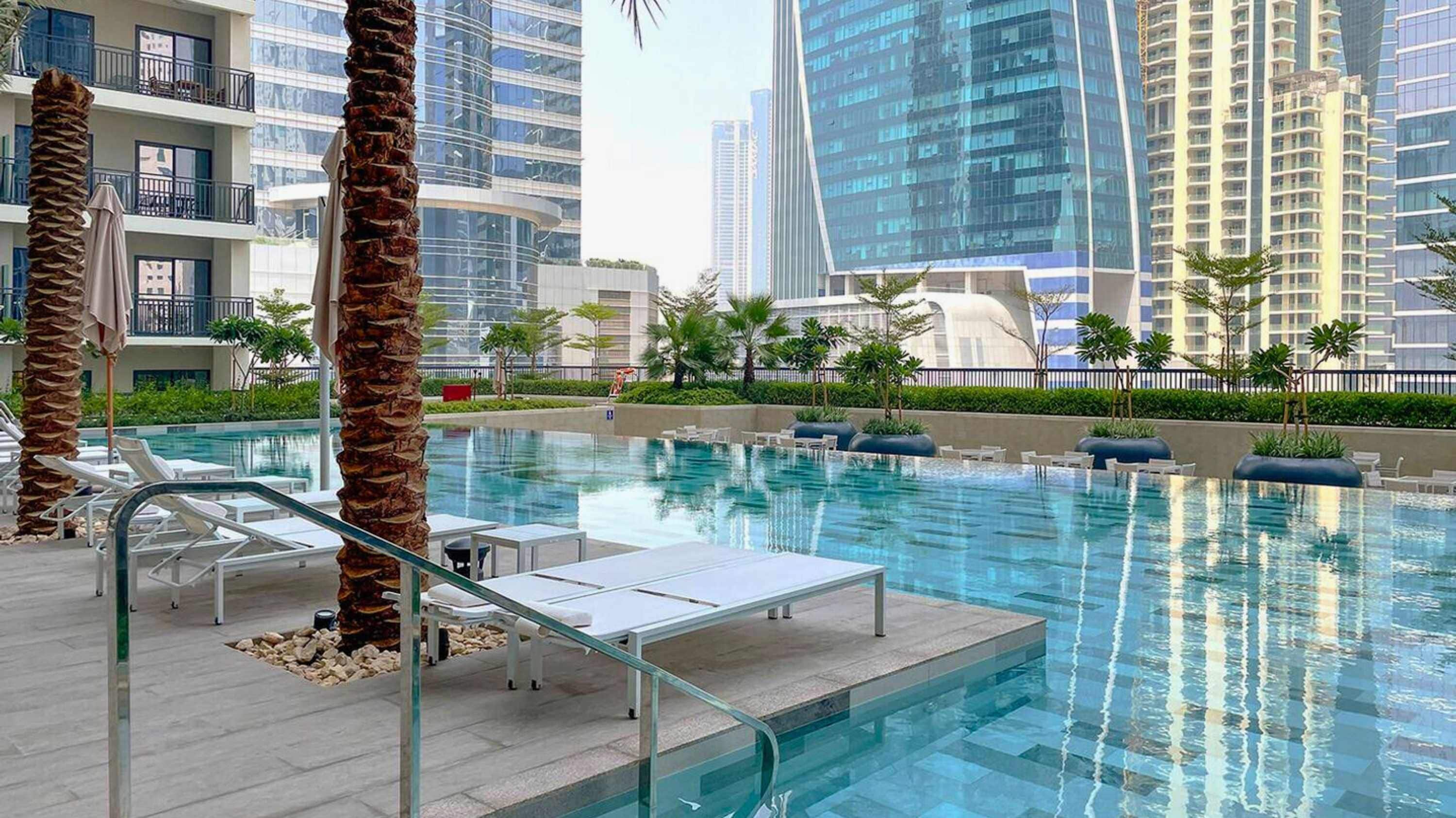 A swimming pool with lounge chairs and palm trees, surrounded by high-rise buildings.