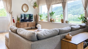 Ein modernes Wohnzimmer mit einem großen grauen Sofa, einem Fernseher auf einer Holzkommode und Blick ins Grüne durch große Fenster.