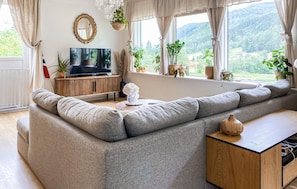A modern living room with a large grey sofa, a television on a wooden cabinet, and a view of greenery through large windows. - 5 bedroom lovely home in Syvde (Syvde)