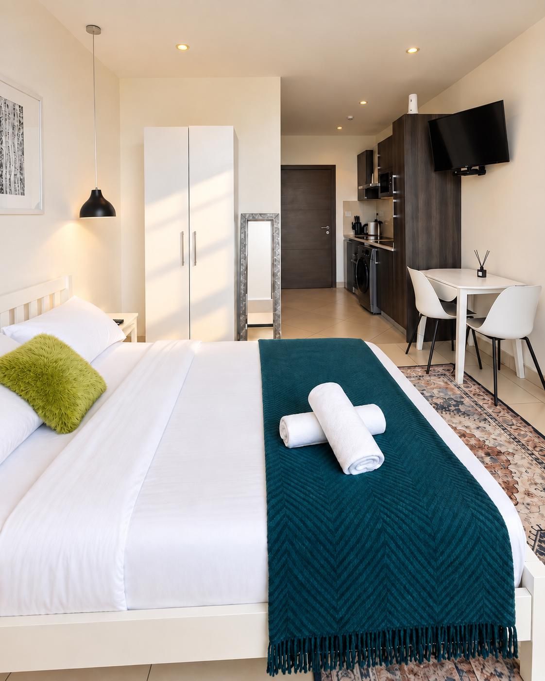 A hotel room with a bed, a desk, a chair, a TV, and a kitchenette.