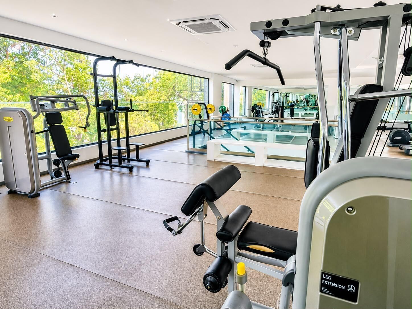 Une salle de sport équipée de divers appareils d’exercice, dotée de grandes fenêtres et offrant une vue sur la verdure environnante.