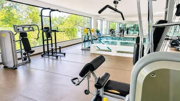 Een sportschool met diverse fitnessapparatuur, grote ramen en uitzicht op groen.