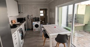 A compact kitchen with a washing machine, a dining table with chairs, and a view of the outside through a glass door. - Azores Green Escape (Povoação)