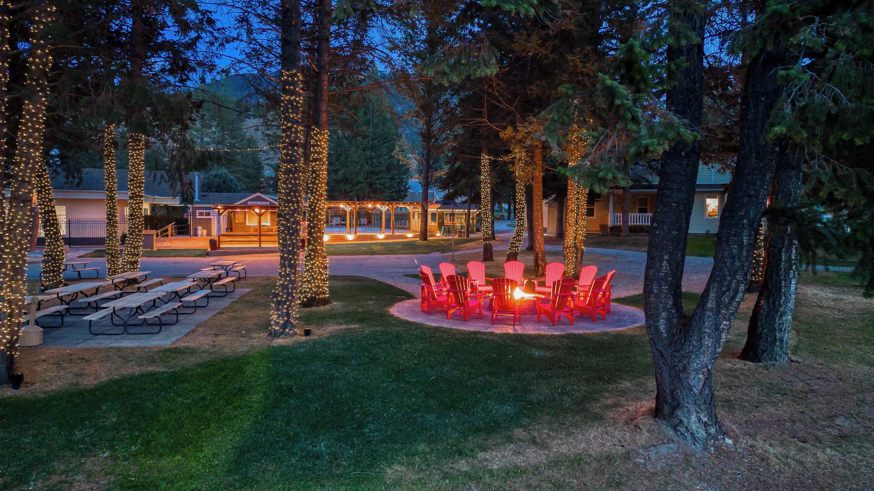 A well-lit outdoor area with picnic tables and red chairs arranged around a fire pit.