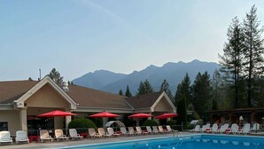 A swimming pool with a metal ladder, lounge chairs, and umbrellas, with mountains in the background. - Mountain Hearth - a RidgeView Resort Partner Property (Radium Hot Springs)