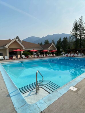 Ein Schwimmbecken mit Metallleiter, Liegestühlen und Sonnenschirmen, im Hintergrund Berge.