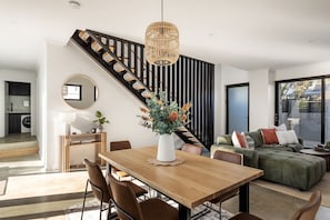 A modern living room with a wooden dining table, a sofa, a staircase, and a large window. - Stone River Villa 4 | Serene Escape | Heated Pool (Margaret River)