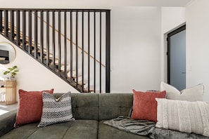 Ein Wohnzimmer mit grauem Sofa, gemusterten Kissen, einer Treppe mit schwarzen Geländern und einer blauen Tür.