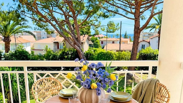 A round table with green plates and a vase of flowers, surrounded by wicker chairs with cushions, on a balcony with a view of houses and trees. - 06. Bright One-Bedroom Apartment with Terrace, Pool, Tennis Court and Garage (Vallauris)