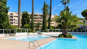 A swimming pool with a clear blue water surface, surrounded by palm trees and a white fence. - 06. Bright One-Bedroom Apartment with Terrace, Pool, Tennis Court and Garage (Vallauris)