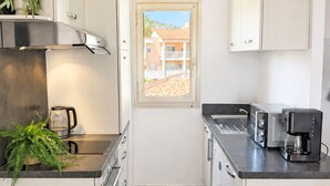 A compact kitchen with light wood cabinets, stainless steel appliances, and a window with a view of buildings. - 06. Bright One-Bedroom Apartment with Terrace, Pool, Tennis Court and Garage (Vallauris)