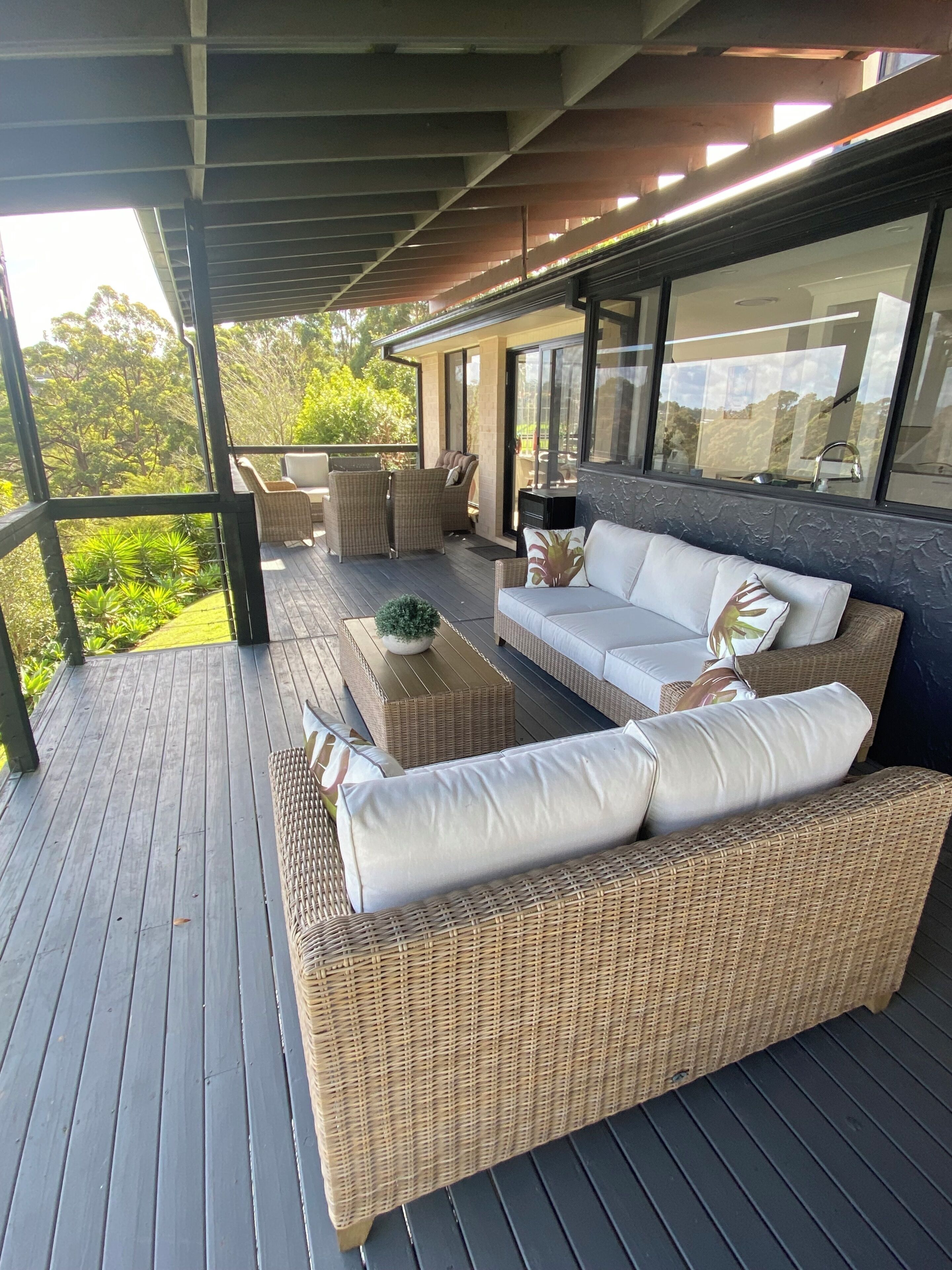 Un espace extérieur aménagé avec des meubles en osier, protégé par une structure en verre, et offrant une vue sur la verdure.
