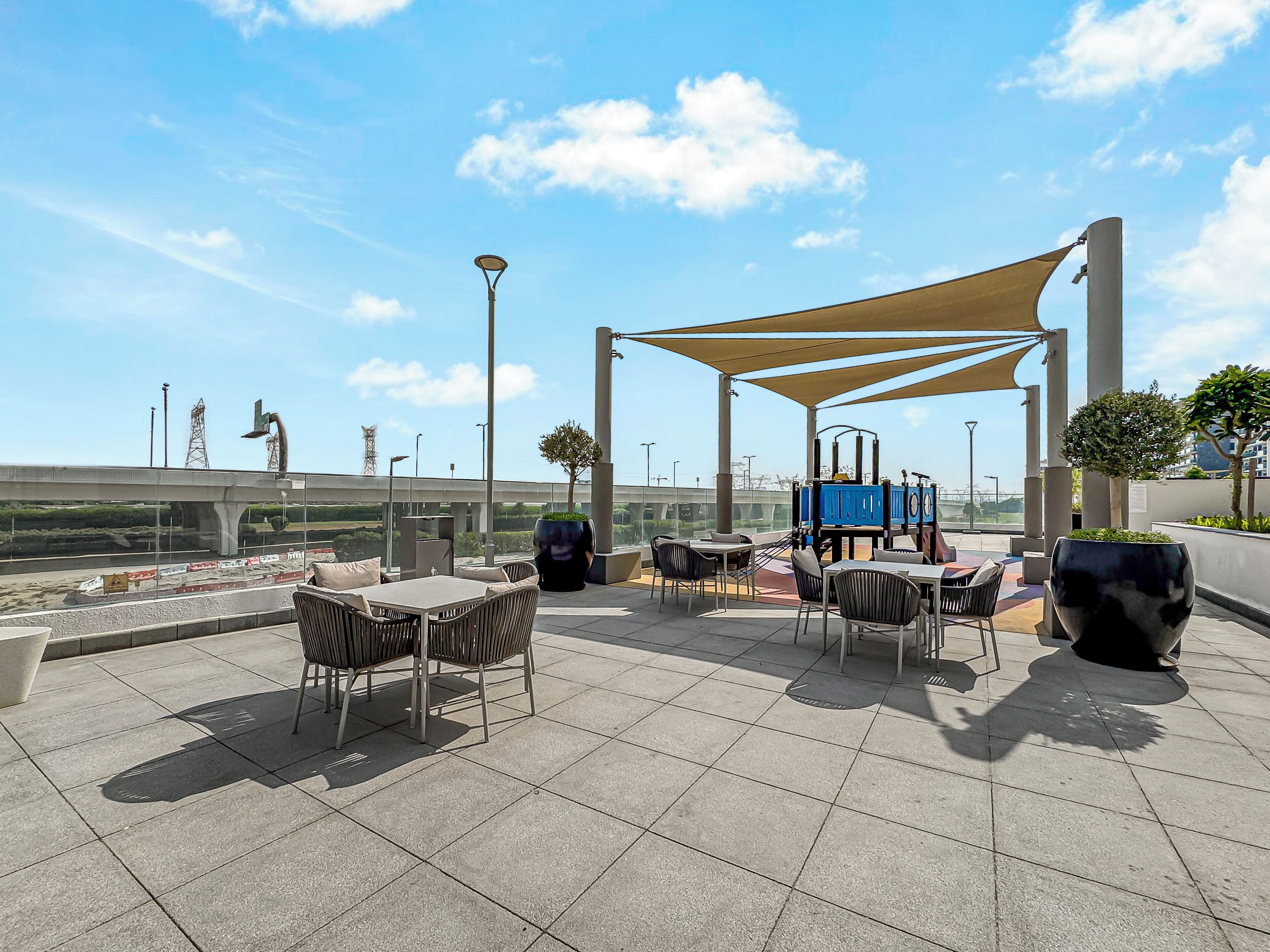 Un patio exterior con mesas y sillas, una estructura de juegos y vistas a un puente y una autopista.