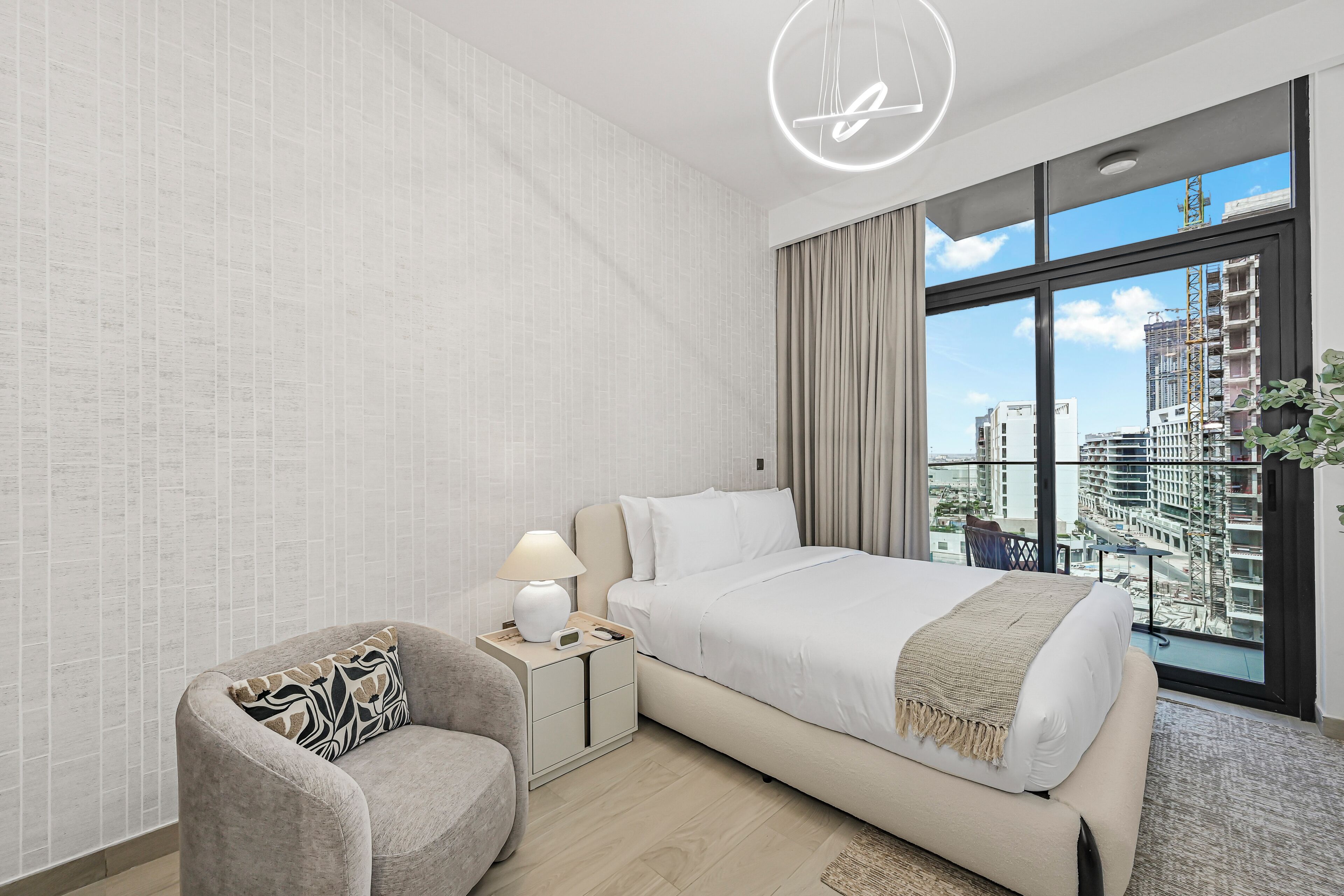 Un dormitorio moderno con una cama grande, una mesita de noche con lámpara, una silla y vistas a la ciudad a través de amplias ventanas.
