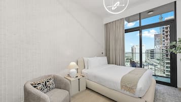 Un dormitorio moderno con una cama grande, una mesita de noche con lámpara, una silla y vistas a la ciudad a través de amplias ventanas.