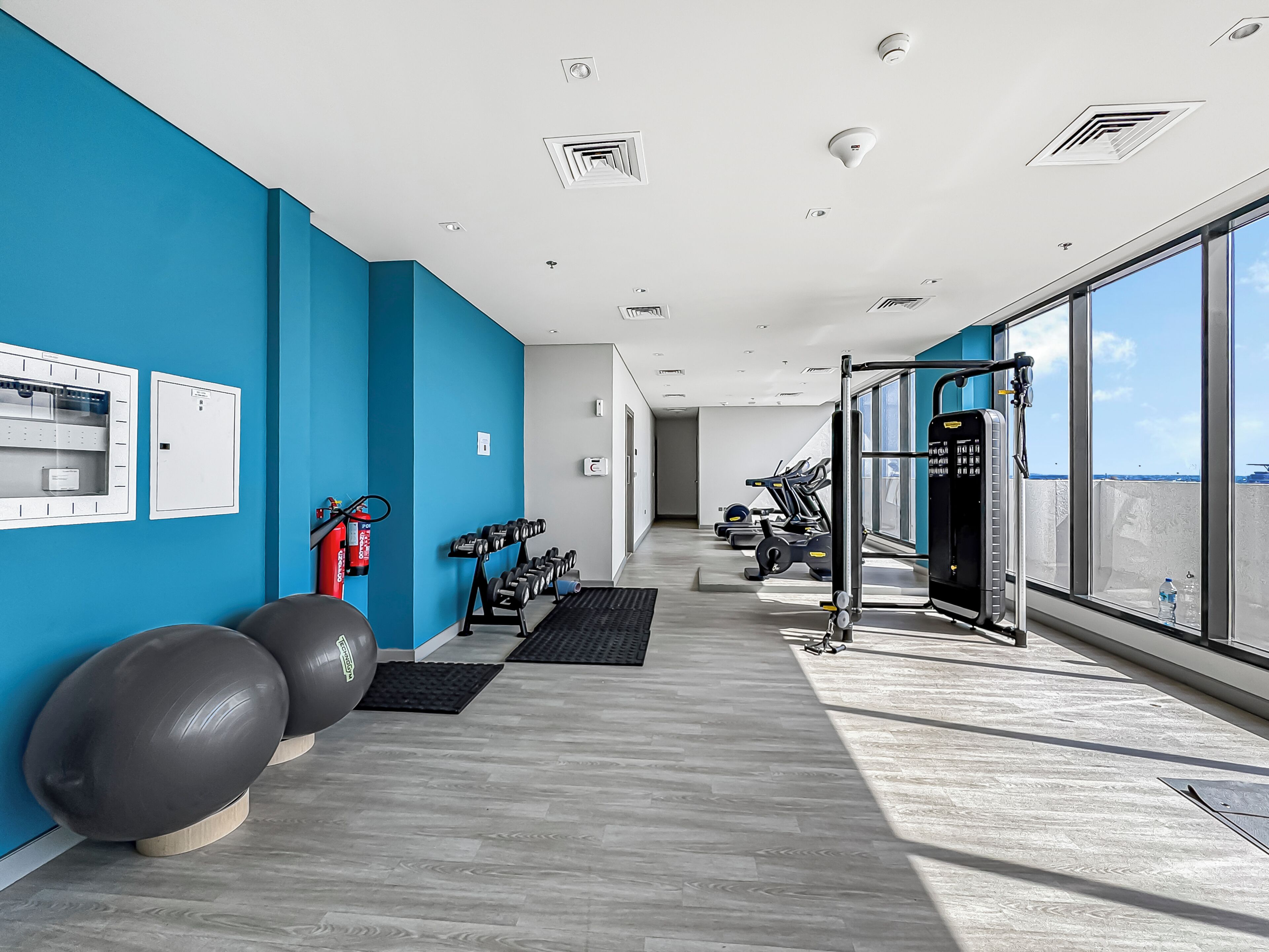 Un gimnasio moderno con equipo de ejercicio, un extintor y vistas al cielo a través de amplias ventanas.