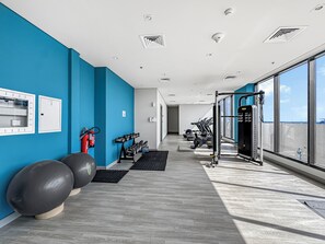 Ein modernes Fitnessstudio mit Trainingsgeräten, einem Feuerlöscher und Blick zum Himmel durch große Fenster.