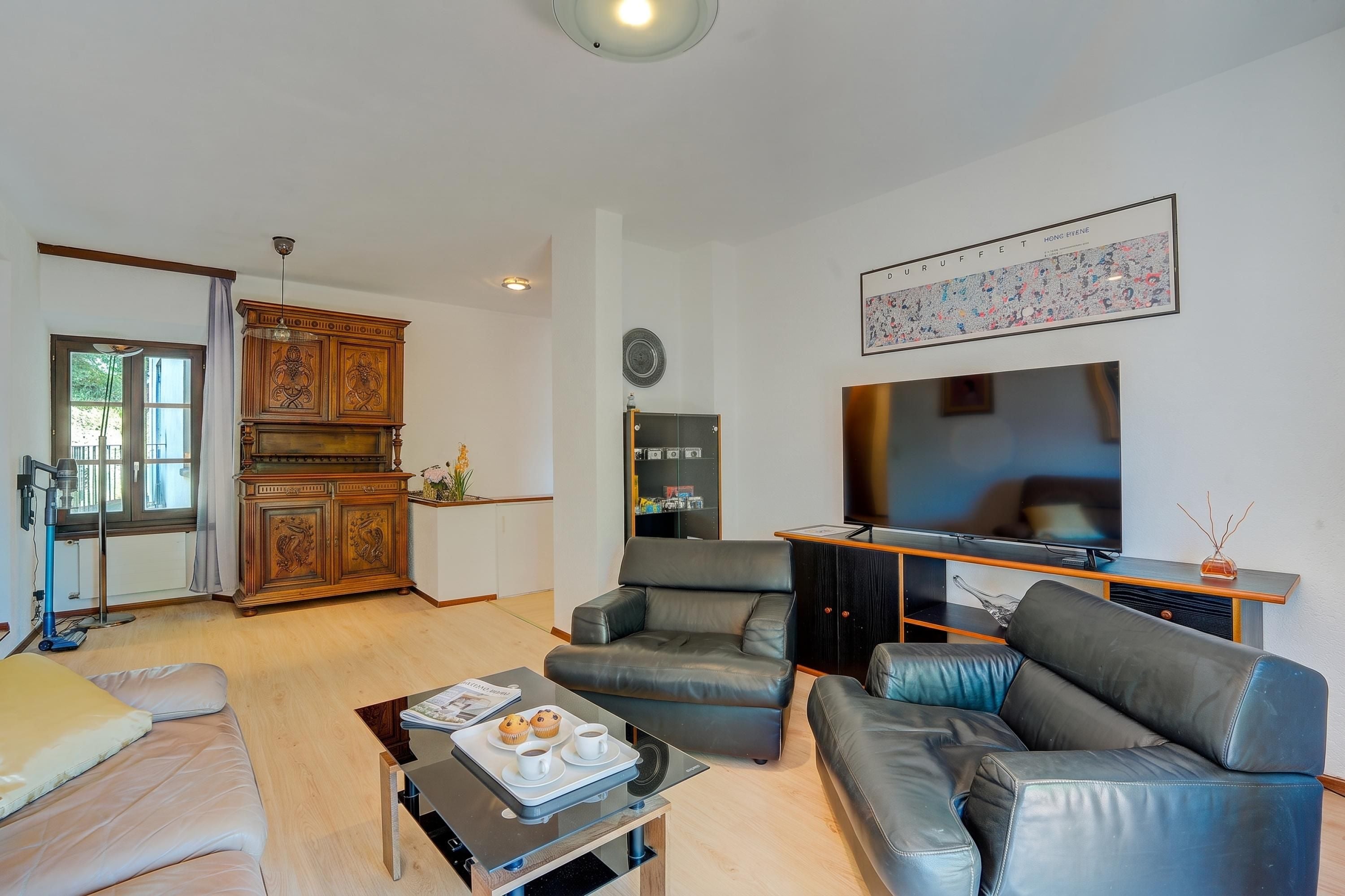 Ein Wohnzimmer mit Sofa, Couchtisch und Fernseher. Es gibt einen Schrank mit dekorativen Schnitzereien und ein Fenster mit Vorhängen.
