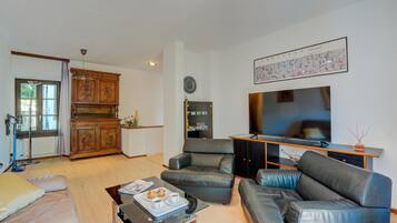 Ein Wohnzimmer mit Sofa, Couchtisch und Fernseher. Es gibt einen Schrank mit dekorativen Schnitzereien und ein Fenster mit Vorhängen.