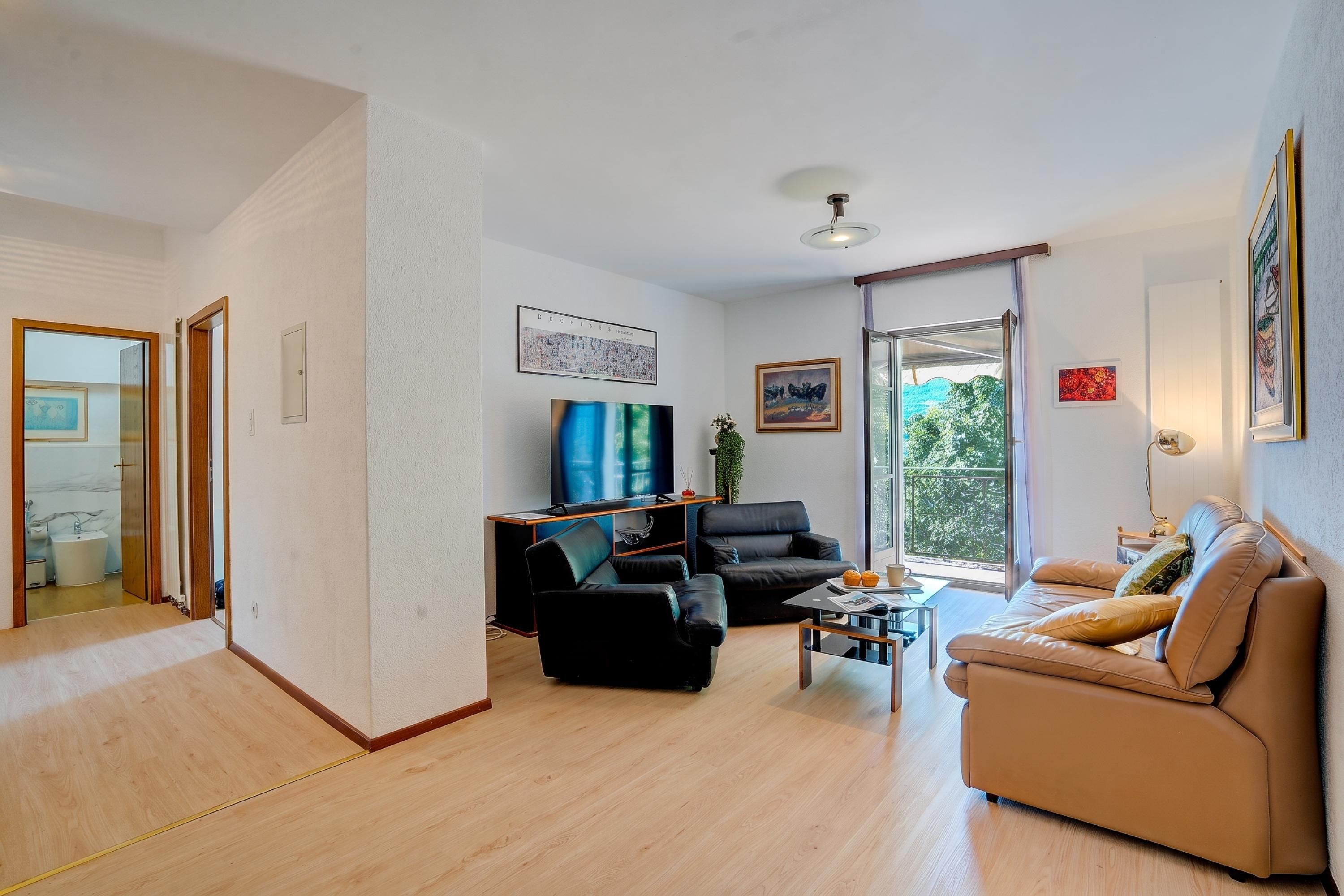 Ein modernes Wohnzimmer mit einer Couch, einem schwarzen Sessel und einem Couchtisch. Ein Fernseher ist an der Wand montiert, und ein Fenster bietet Blick auf Grünflächen.