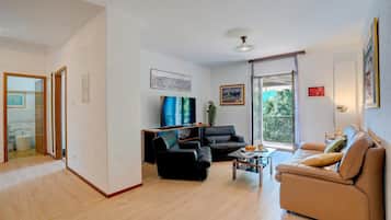 Ein modernes Wohnzimmer mit einer Couch, einem schwarzen Sessel und einem Couchtisch. Ein Fernseher ist an der Wand montiert, und ein Fenster bietet Blick auf Grünflächen.