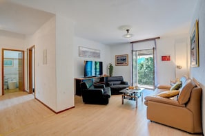 A modern living room with a sofa, a black armchair, and a coffee table. There is a television mounted on the wall, and a window with a view of greenery outside. - Casa Pontaccio With Terrace And View (Lugano)
