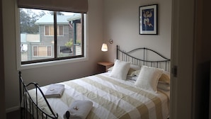 Ein Schlafzimmer mit einem Bett, einem Nachttisch, einer Wandlampe, einem Deckenventilator und einem Fenster mit Blick auf ein Haus.