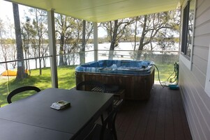 A covered patio with a hot tub, a table, and chairs, overlooking a body of water and trees.