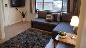 Ein Wohnzimmer mit einer Couch, einem Flachbildfernseher, einem Topf mit Pflanze und einem Fenster mit Vorhängen.