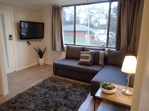 A living room with a sofa, a flat-screen TV, a potted plant, and a window with curtains. - Dungowan Holiday Accomodation (Sanctuary Point)