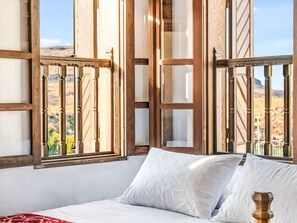 Een slaapkamer met een bed, witte kussens en houten raamkozijnen met uitzicht op een woestijnlandschap.