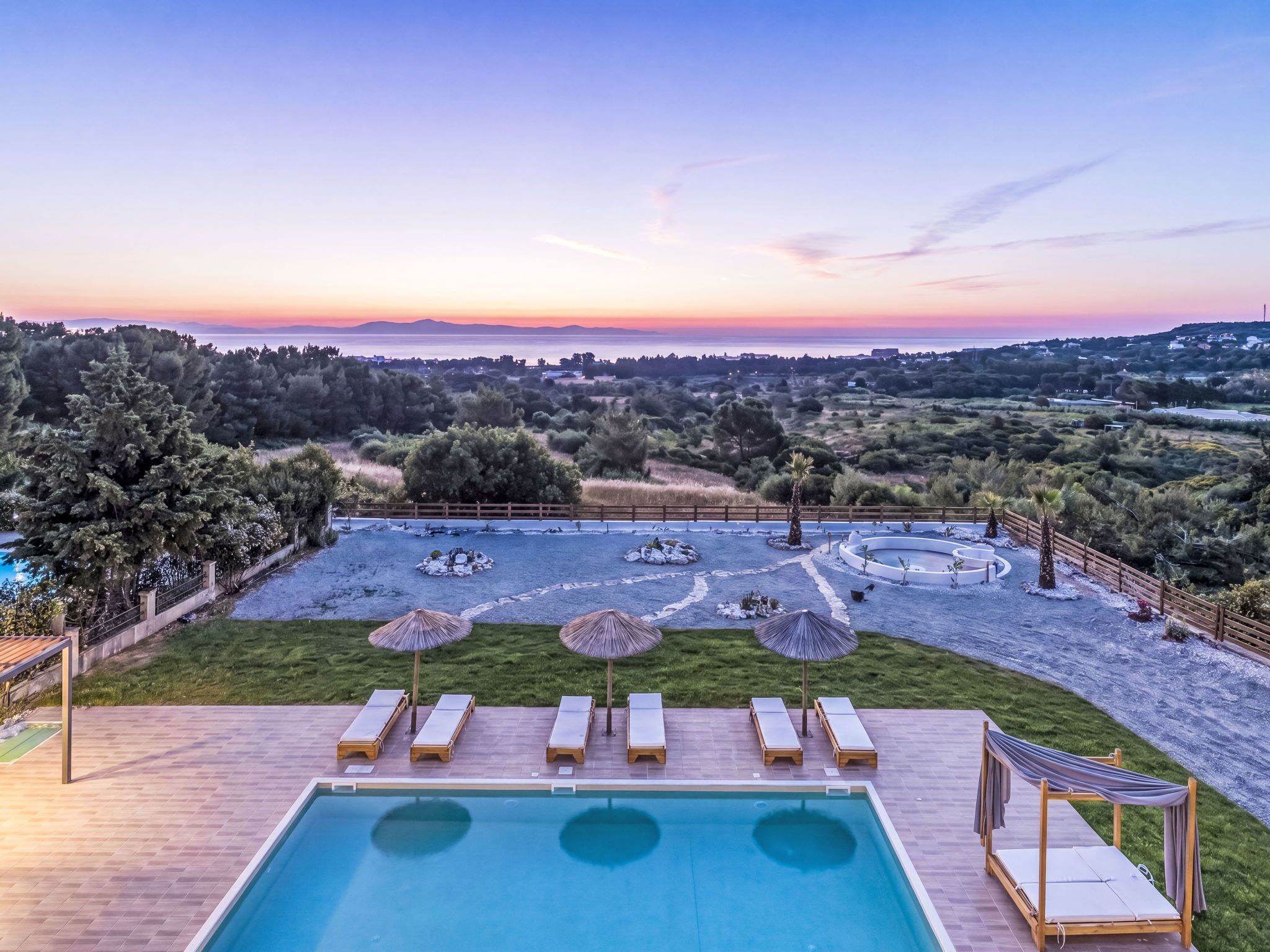Une piscine entourée d’un espace aménagé avec des arbres, des chaises longues et des parasols, offrant une vue sur le paysage.