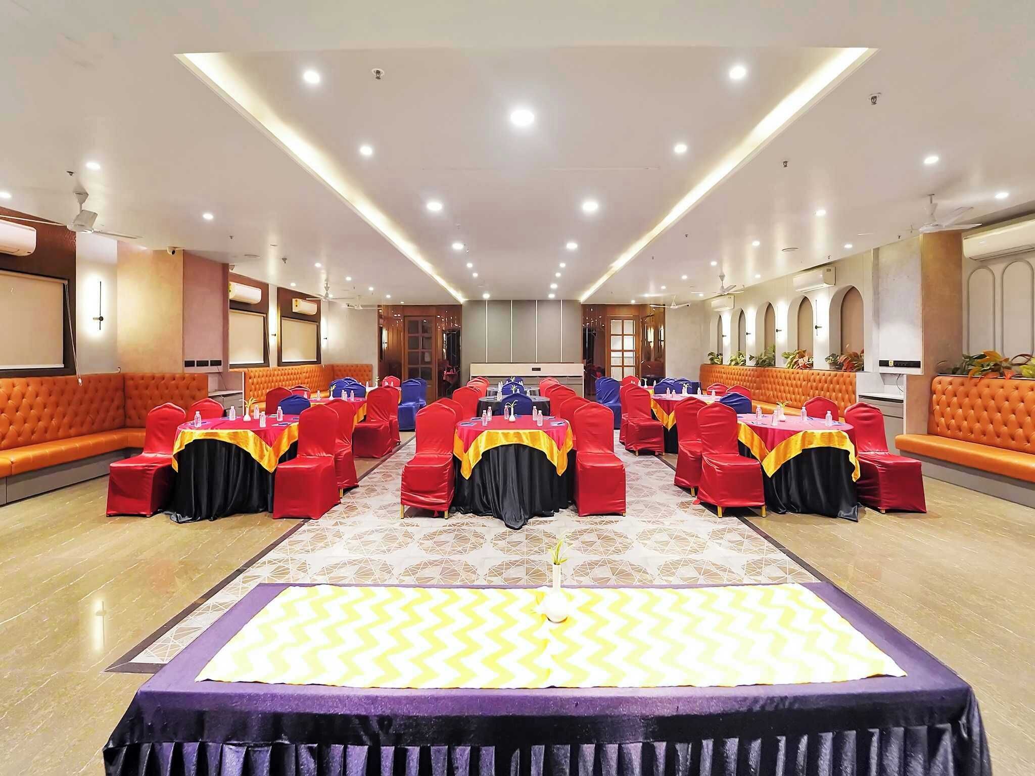 A conference room with rows of red chairs, a central stage with a purple and yellow carpet, and a modern ceiling design.