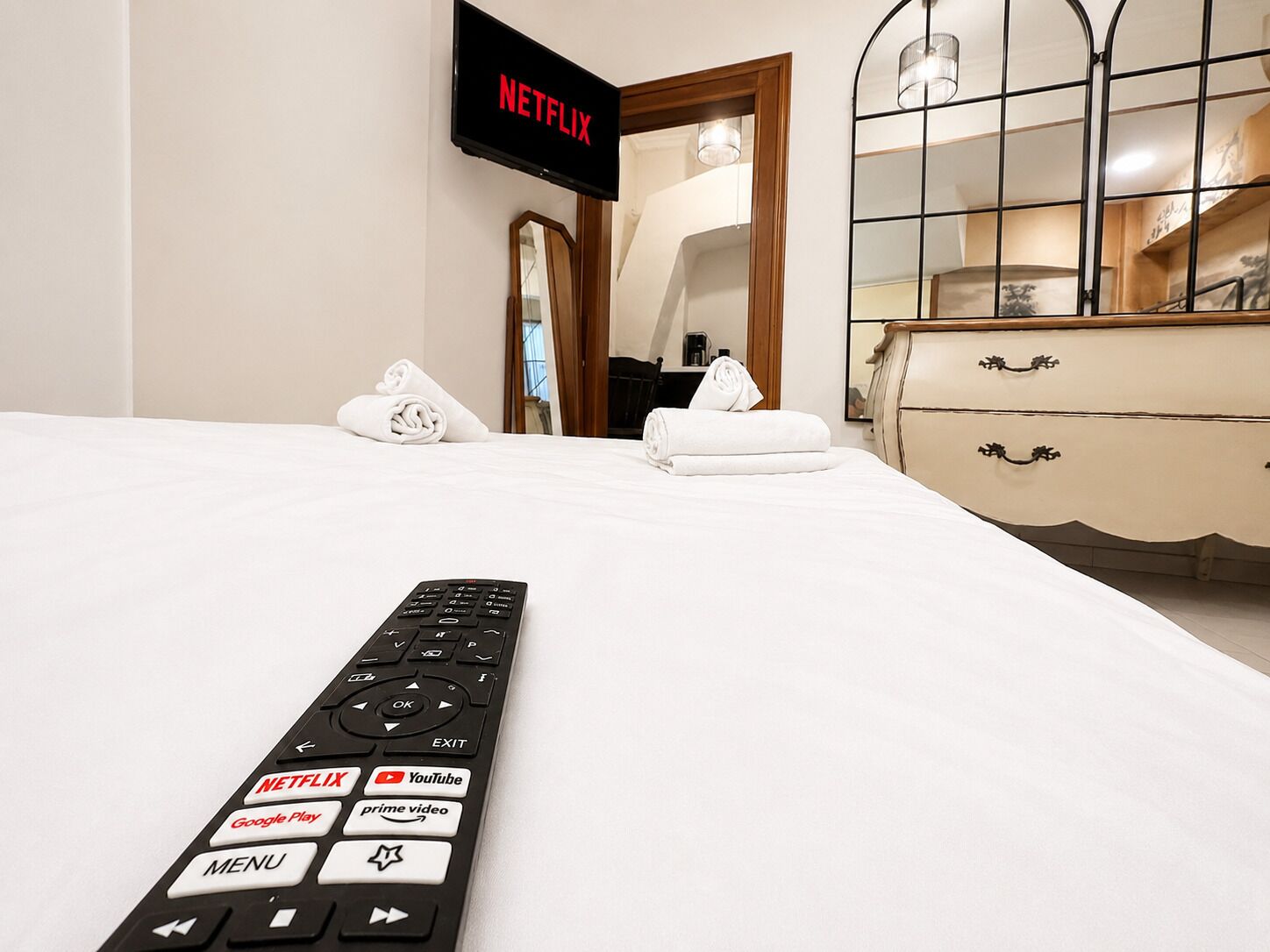 A hotel room with a bed, a remote control with Netflix and YouTube buttons, a TV displaying 'NETFLIX', and a dresser with drawers.