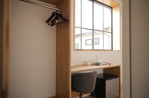 Una habitación con un escritorio de madera, una silla, un pequeño electrodoméstico negro y una ventana con vista a edificios.