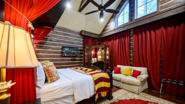 A bedroom with a bed, a sofa, a TV, a ceiling fan, and a large window. - Themed Cabin+Treehouse | 5 King Suites | Sleeps 23 (Broken Bow)