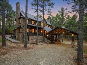 A log cabin with a stone chimney, a covered porch, and a gravel driveway surrounded by trees. - Wizard Cabin+Treehouse | Game Zone | Sleeps 23 (Broken Bow)