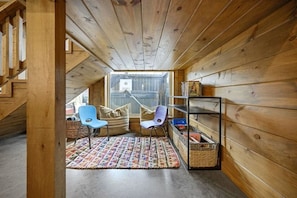 Ein gemütliches Dachzimmer mit Holzwänden, schrägen Decken, zwei Stühlen, einem kleinen Regal und Blick durch ein Fenster.