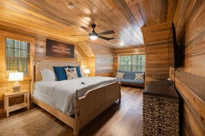 A cozy wooden cabin bedroom with a bed, a bench, a ceiling fan, and a mounted TV. - 2 King Suites | Hot Tub+Game Room | Sleeps 8 (Broken Bow)
