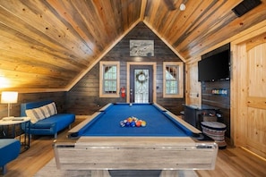 A pool table with balls, a blue sofa, and a fireplace in a wooden room with a sloped ceiling. - 2 King Suites | Hot Tub+Game Room | Sleeps 8 (Broken Bow)