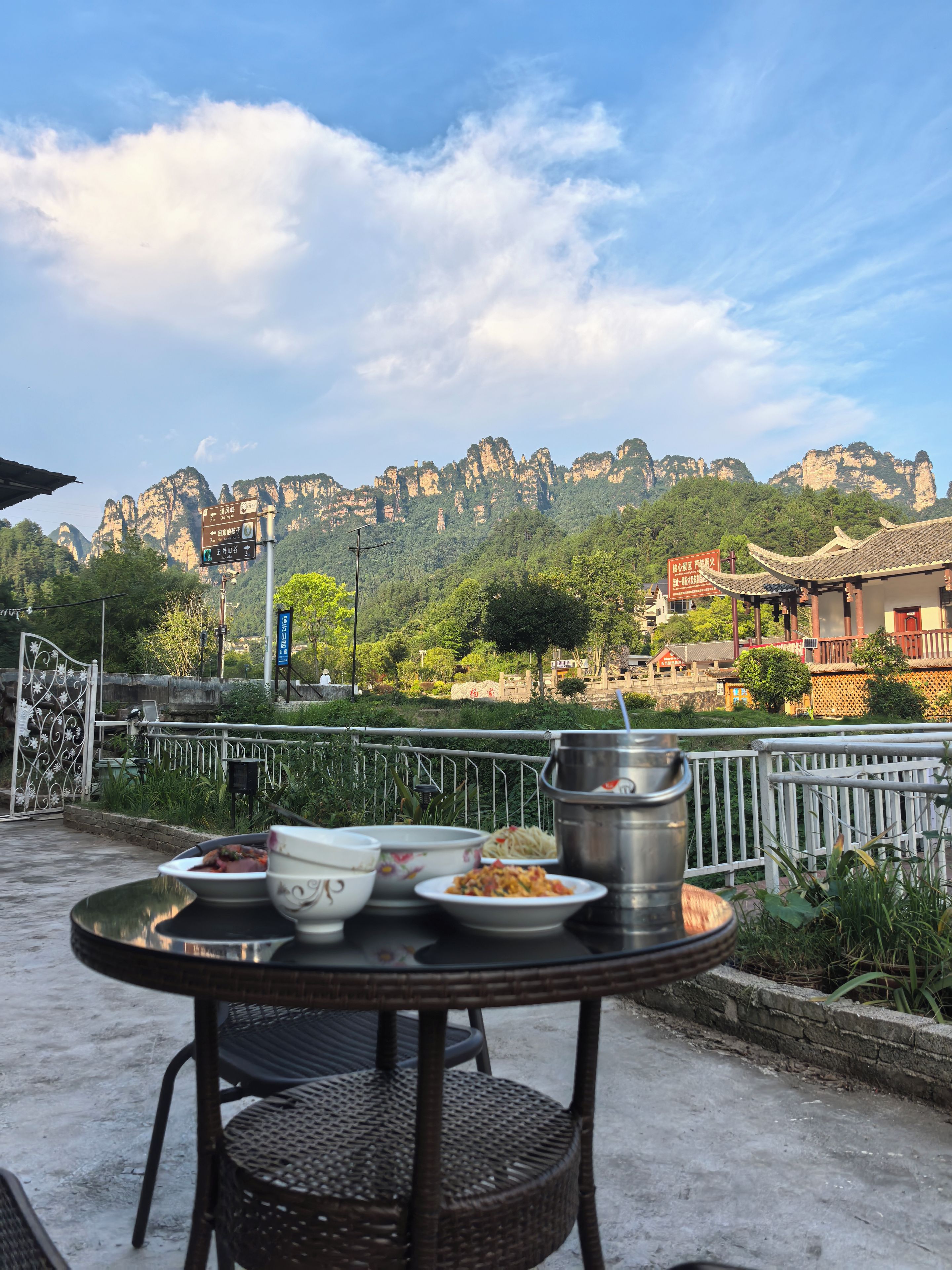 Una mesa con platos y un recipiente metálico, con montañas y arquitectura tradicional como telón de fondo.