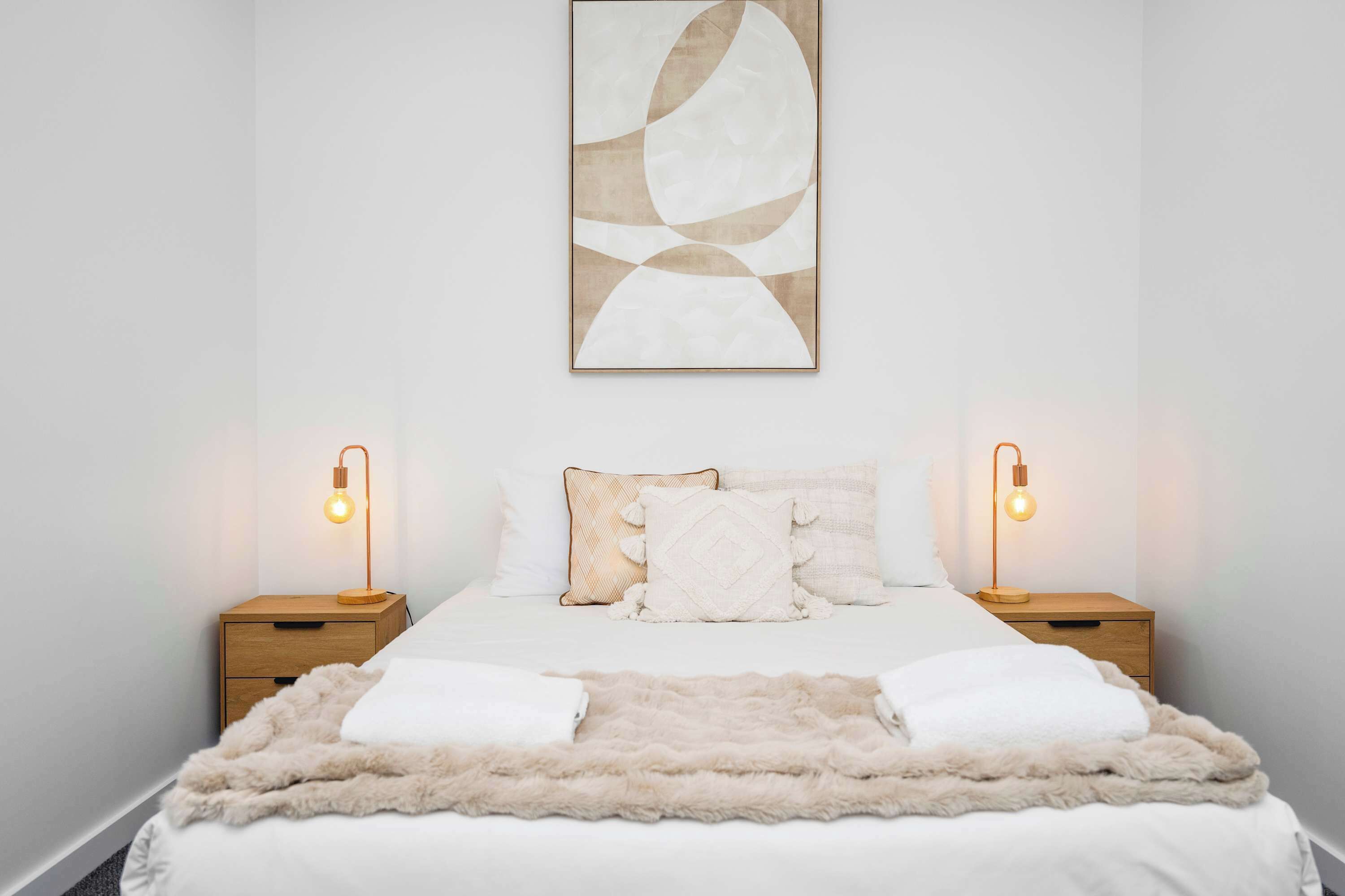 Un dormitorio ordenado con una cama, dos mesitas de noche, un tapete mullido y una obra de arte abstracto colgada en la pared.
