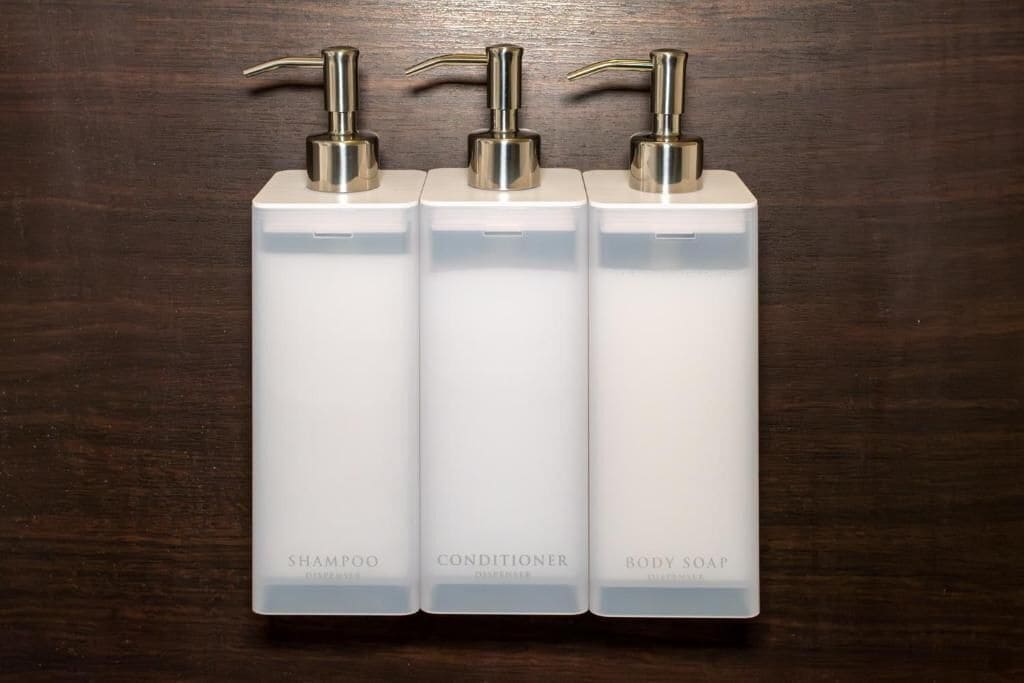 Three white bottles with pumps, labeled 'SHAMPOO,' 'CONDITIONER,' and 'BODY SOAP,' on a wooden surface.