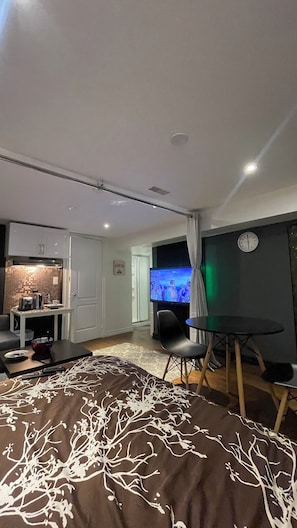 Ein modernes Wohnzimmer mit Flachbildfernseher, Esstisch und einem gemütlichen Bett mit gemustertem Überwurf.