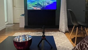 A living room with a flat-screen TV mounted on a dark wall, a tripod with a camera on a table, and a chair.